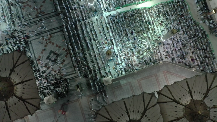 An aerial view of the Prophet's Mosque in Medina at night, showing the worshippers and umbrellas.