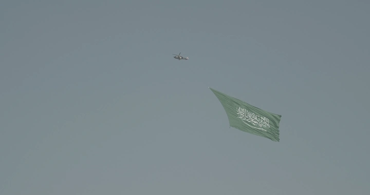Helicopter carrying Saudi flag in the sky, daytime shot.
