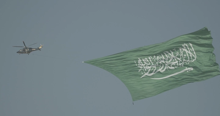 Helicopter carrying Saudi flag in air show, daytime shot.