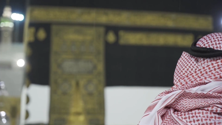 رجل يرتدي شماغًا أمام الكعبة في الليل، لقطة مقربة.