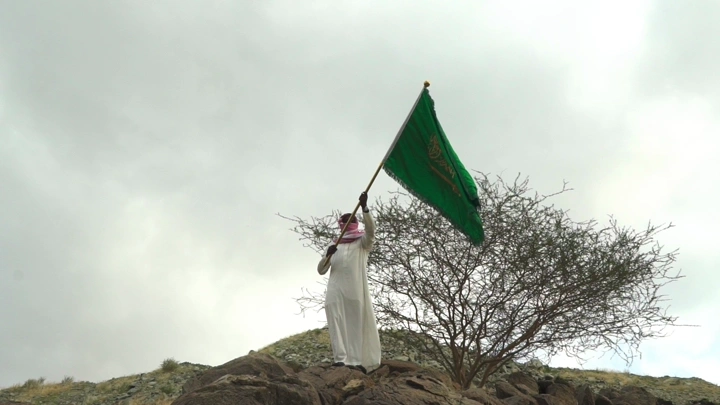 رجل يحمل علمًا أخضر على قمة جبل تحت سماء غائمة، لقطة من الأسفل.