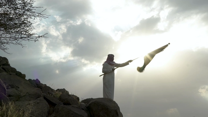 شخص يرفع علمًا على تل صخري عند غروب الشمس، لقطة جانبية.