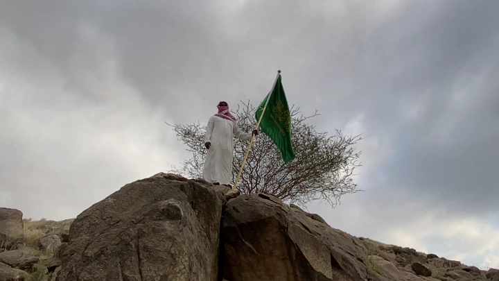 شخص يحمل علمًا على صخرة في يوم غائم، لقطة من الأرض.