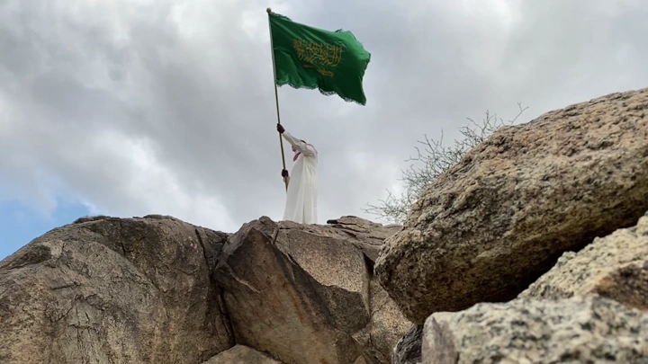 شخص يرفع علمًا أخضر على قمة جبلية، لقطة من الأسفل.