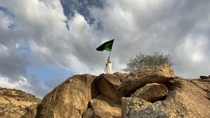 شخص يحمل علمًا على قمة جبل تحت سماء غائمة، لقطة من الأسفل.