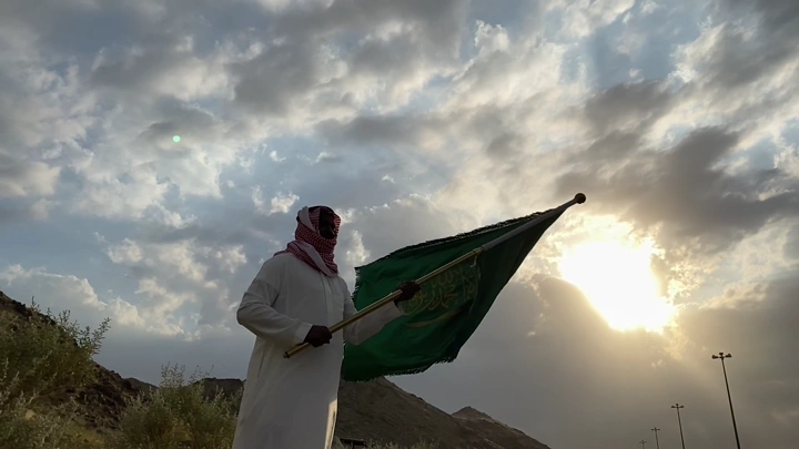 رجل يحمل علمًا في منطقة جبلية عند غروب الشمس، لقطة من زاوية منخفضة.