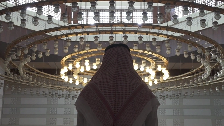 شخص يرتدي ثوبًا تقليديًا في مسجد تحت ثريا ضخمة، لقطة داخلية.