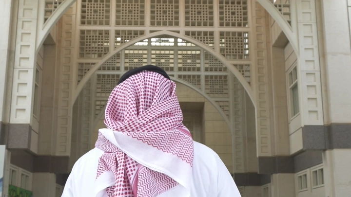 رجل يرتدي شماغ أمام بوابة جامعة، لقطة من الخلف.