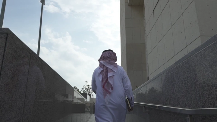 طالب يمشي في ممر خارجي بجانب مبنى حديث، لقطة من الخلف.