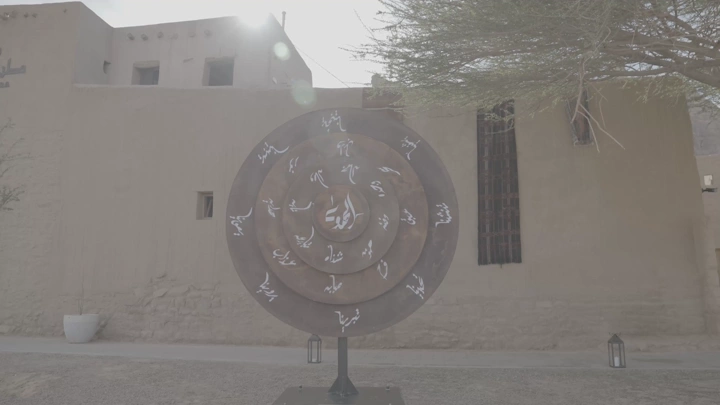تمثال دائري مع كتابات عربية أمام مبنى تقليدي في العلا، لقطة نهارية.