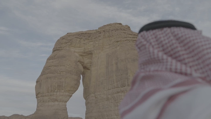 رجل يرتدي شماغًا ينظر إلى صخرة الفيل في العلا، لقطة من الخلف.