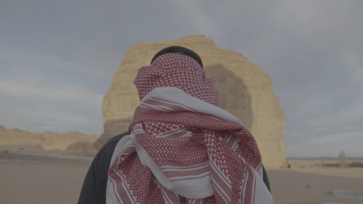 رجل يرتدي شماغًا أمام صخرة في العلا، لقطة خلفية.