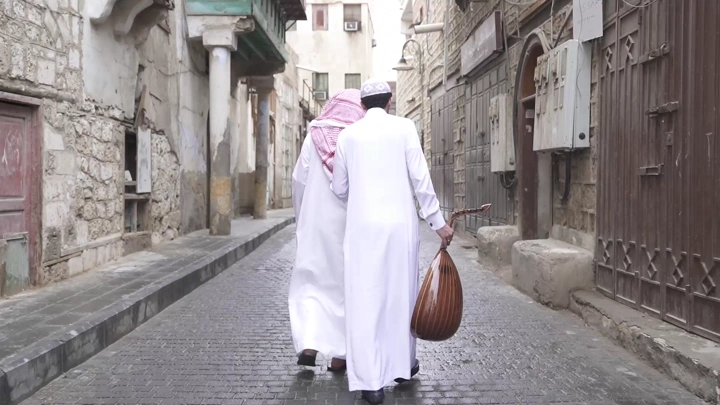 شاب سعودي يساعد شخص كفيف في شارع تاريخي، لقطة من الخلف.