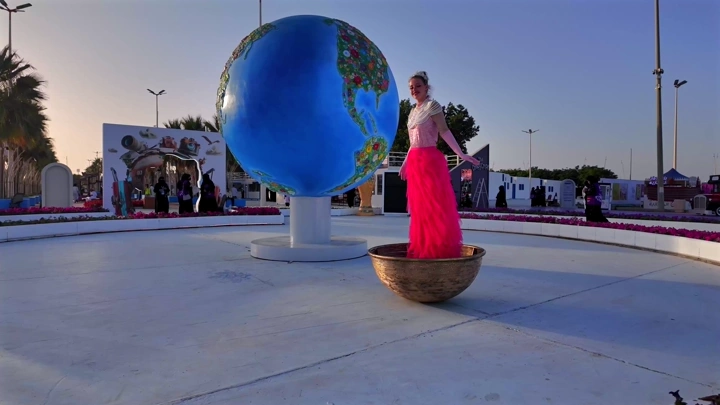 A woman stands next to a globe sculpture at the Flowers and Gardens Festival in Yanbu Industrial City, daytime shot.