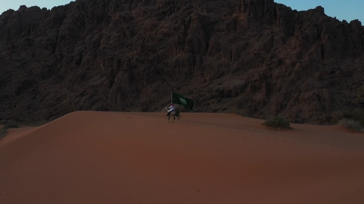 شخص بالزي السعودي يحمل علم السعودية في الصحراء أمام الجبال، لقطة جوية.