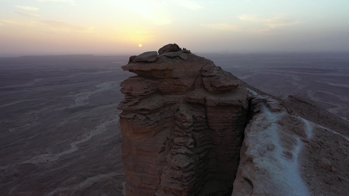 مجموعة اشخاص يقفون فوق جبل طويق عند الغروب، لقطة جوية.