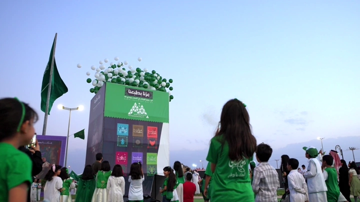 أطفال يرتدون الزي السعودي مع بالونات في احتفال خارجي، لقطة نهارية.