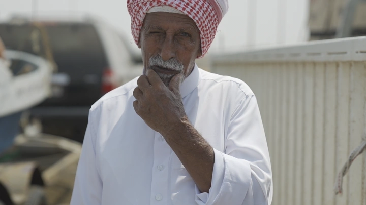 صياد يرتدي غترة وعقال يقف بجانب قارب، لقطة نهارية.