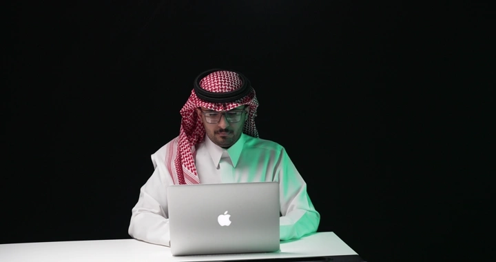 رجل سعودي يعمل على جهاز كمبيوتر محمول في استوديو مظلم.