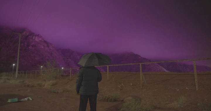 رجل يحمل مظلة أمام جبال حائل تحت سماء بنفسجية، تصوير ليلي.