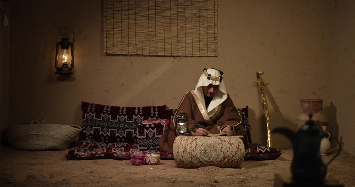 رجل سعودي يكتب في غرفة تقليدية، إضاءة خافتة.