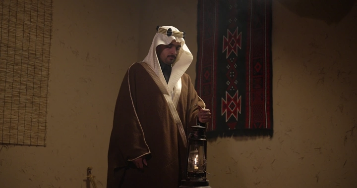 رجل سعودي يرتدي ثوب تقليدي ويمسك سراج في غرفة تقليدية، إضاءة دافئة.