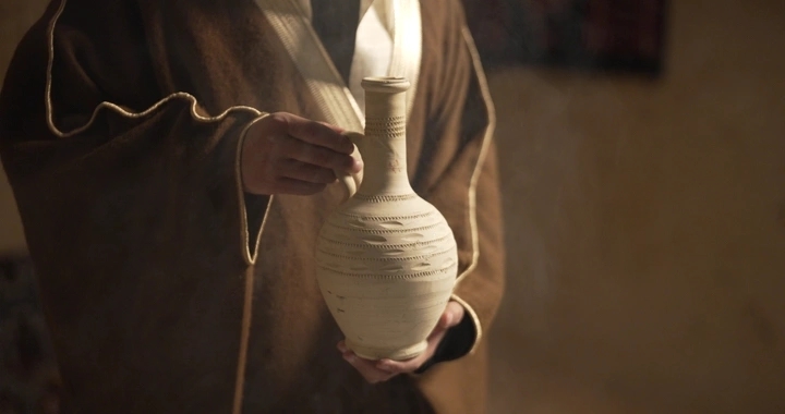 رجل يرتدي ثوبًا تقليديًا يحمل فخارًا، إضاءة دافئة.