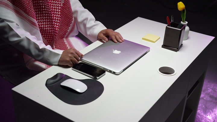 رجل سعودي في مكتب حديث يعمل على لابتوب، لقطة داخلية.