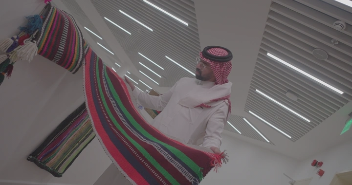 رجل يرتدي الزي التقليدي يحمل سجادة سدو في غرفة حديثة، لقطة داخلية.