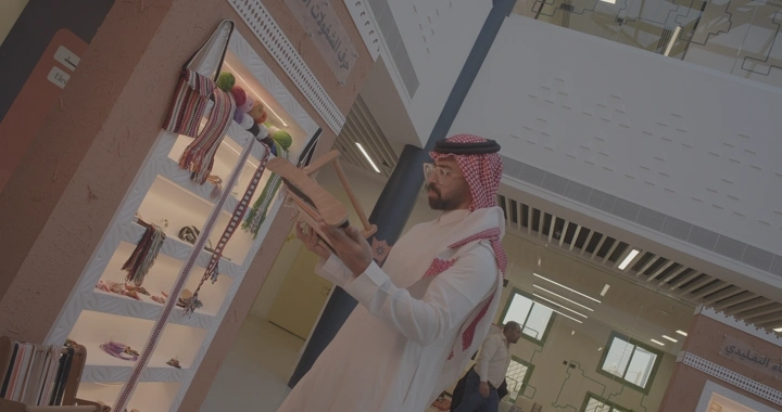 شخص يرتدي الزي السعودي يتأمل أداة نول لصناعة السدو في معرض داخلي.