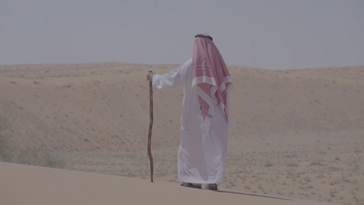 رجل يقف على حفة كثبان رملي و يرتدي ثوبًا وغترة يقف على كثبان رملية في النفود، لقطة نهارية.