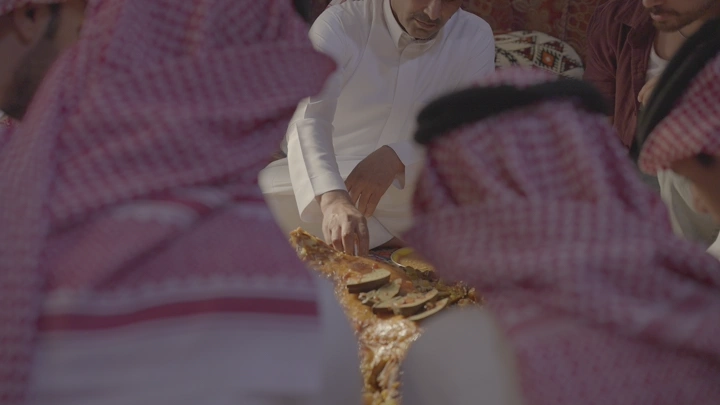 رجال يرتدون الثوب والغترة يجتمعون حول مفطح لحم، لقطة قريبة.