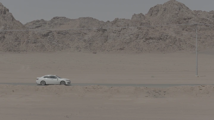 سيارة بيضاء على طريق صحراوي مع جبال في الخلفية، لقطة جانبية.
