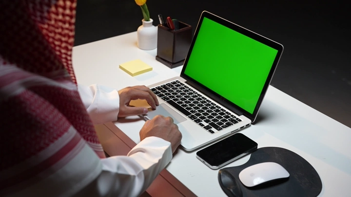 رجل سعودي يعمل على لابتوب مع وجود شاشة خضراء كروما داخل مكتب، لقطة داخلية.