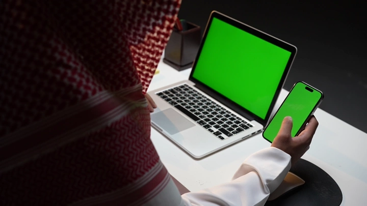 رجل سعودي يعمل على لابتوب وهاتف بشاشة خضراء كروما في مكتب.