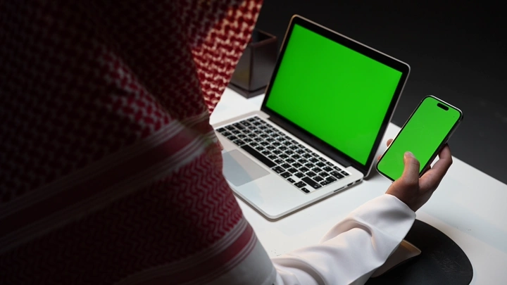 رجل سعودي يعمل على لابتوب وهاتف بشاشة خضراء كروما في مكتب، لقطة قريبة.