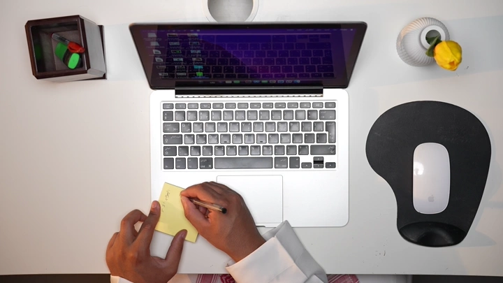 رجل سعودي يعمل على لابتوب ويسجل ملاحظاته في مكتب، لقطة علوية.