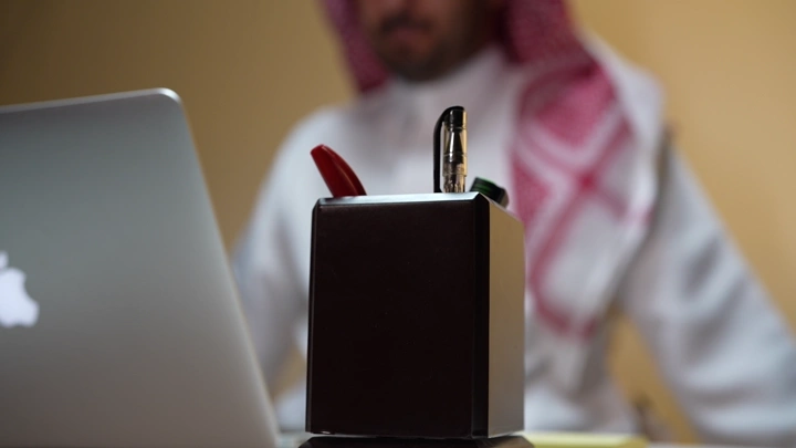 رجل سعودي يعمل على لابتوب في مكتب ويسجل ملاحظاته، لقطة قريبة.