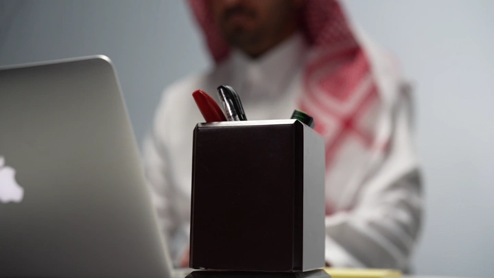 رجل سعودي يعمل على لابتوب ويأخذ قلم من المقلمة داخل مكتب، لقطة مقربة.