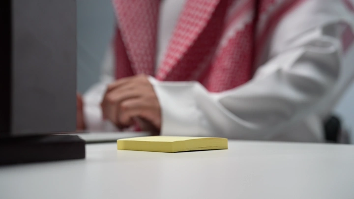 A Saudi man working on a laptop inside an office, taking notes, close-up shot.