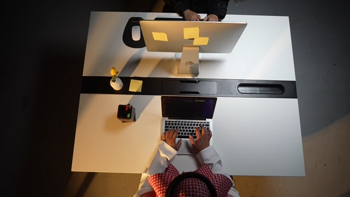 رجل سعودي يعمل على لابتوب في مكتب مع وجود شاشة، لقطة علوية.