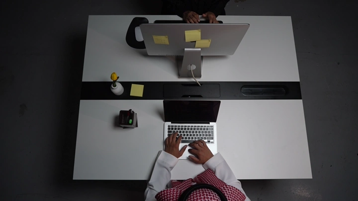 رجل سعودي يعمل على لابتوب في مكتب مع وجود شاشة اخرى، لقطة من الأعلى.