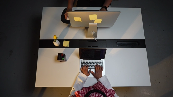 رجل سعودي يعمل على لابتوب في مكتب مع وجود شاشة، لقطة من الأعلى.