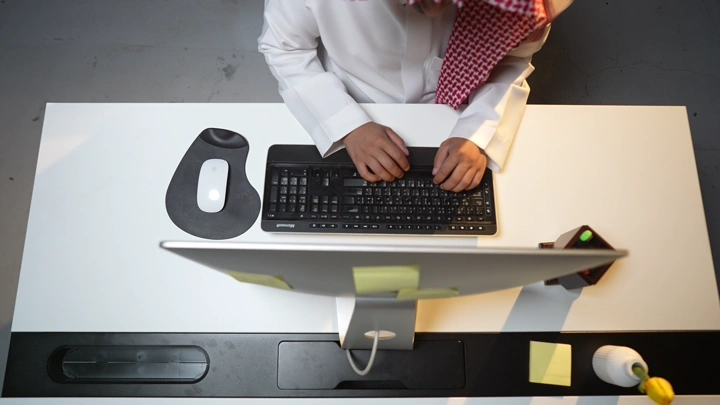 رجل سعودي يعمل على الكمبيوتر في مكتب حديث، لقطة من الأعلى.