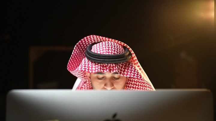 رجل سعودي يعمل على لابتوب في مكتب، إضاءة دافئة.