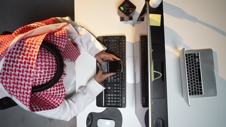 رجل سعودي يعمل على كمبيوتر في مكتب حديث، لقطة علوية.