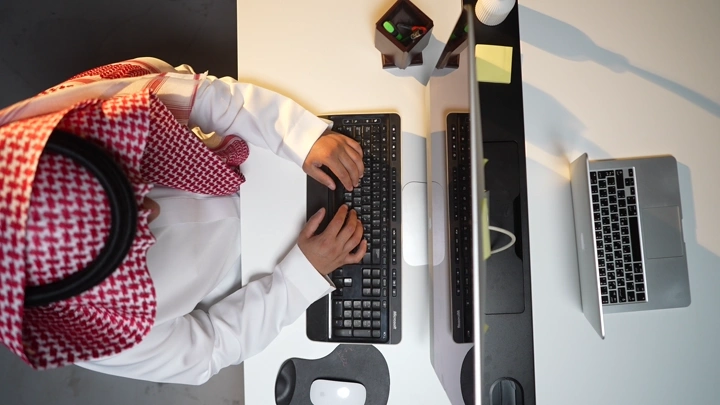 رجل سعودي يعمل على كمبيوتر داخل مكتب، لقطة من الأعلى.