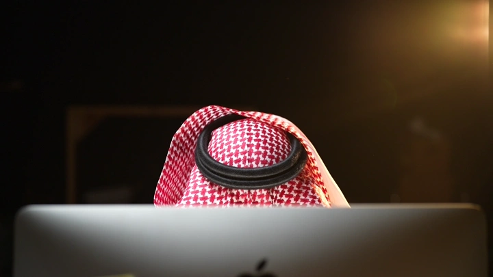 رجل يرتدي شماغ يعمل على كمبيوتر في مكتب، إضاءة خافتة.