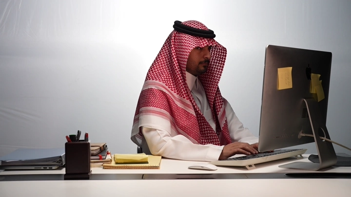 رجل سعودي يعمل على الكمبيوتر في مكتب حديث، لقطة داخلية.