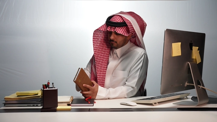 رجل سعودي يقرأ كتابًا في مكتب حديث، لقطة داخلية.